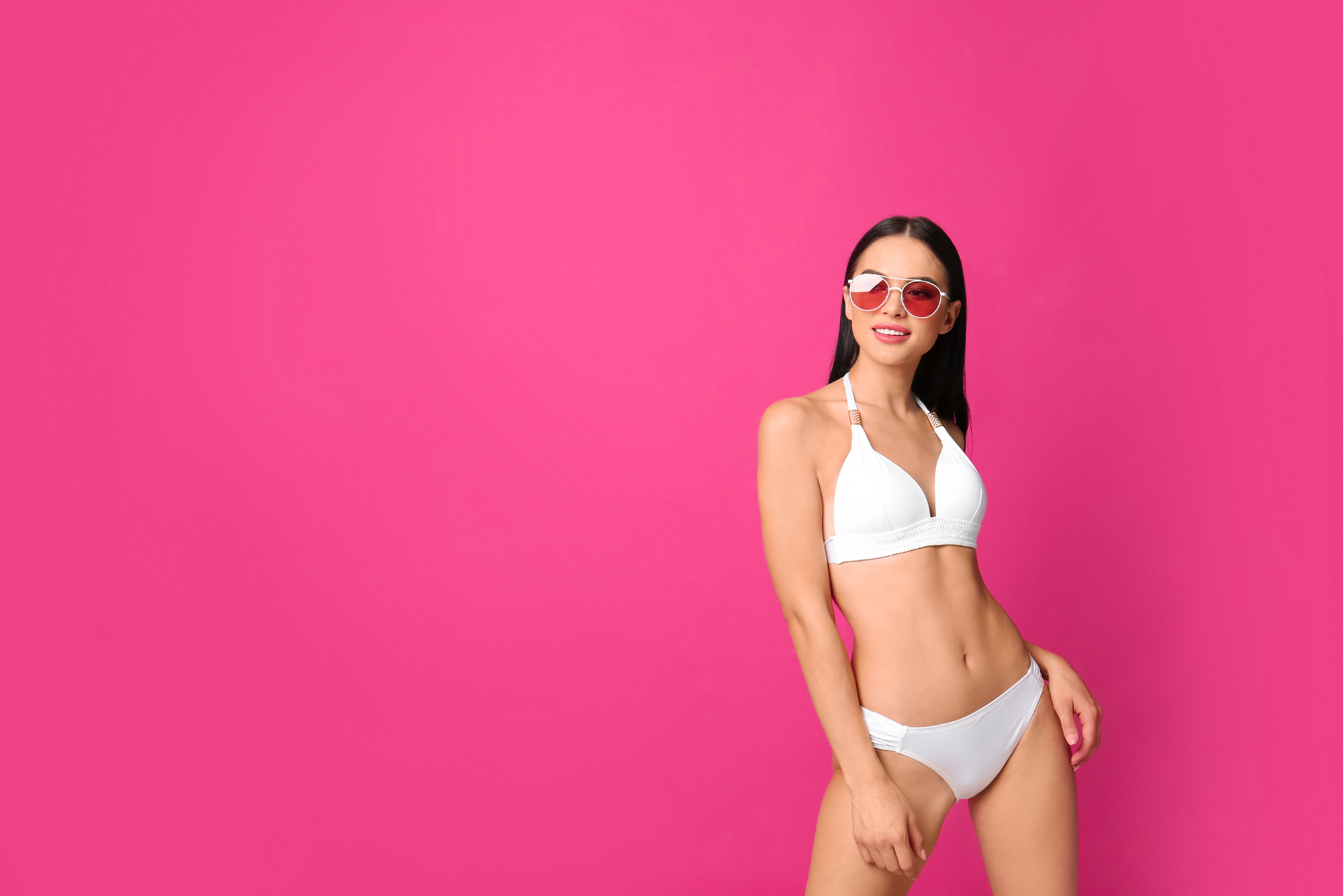 Beautiful young woman in white bikini with sunglasses on pink ba